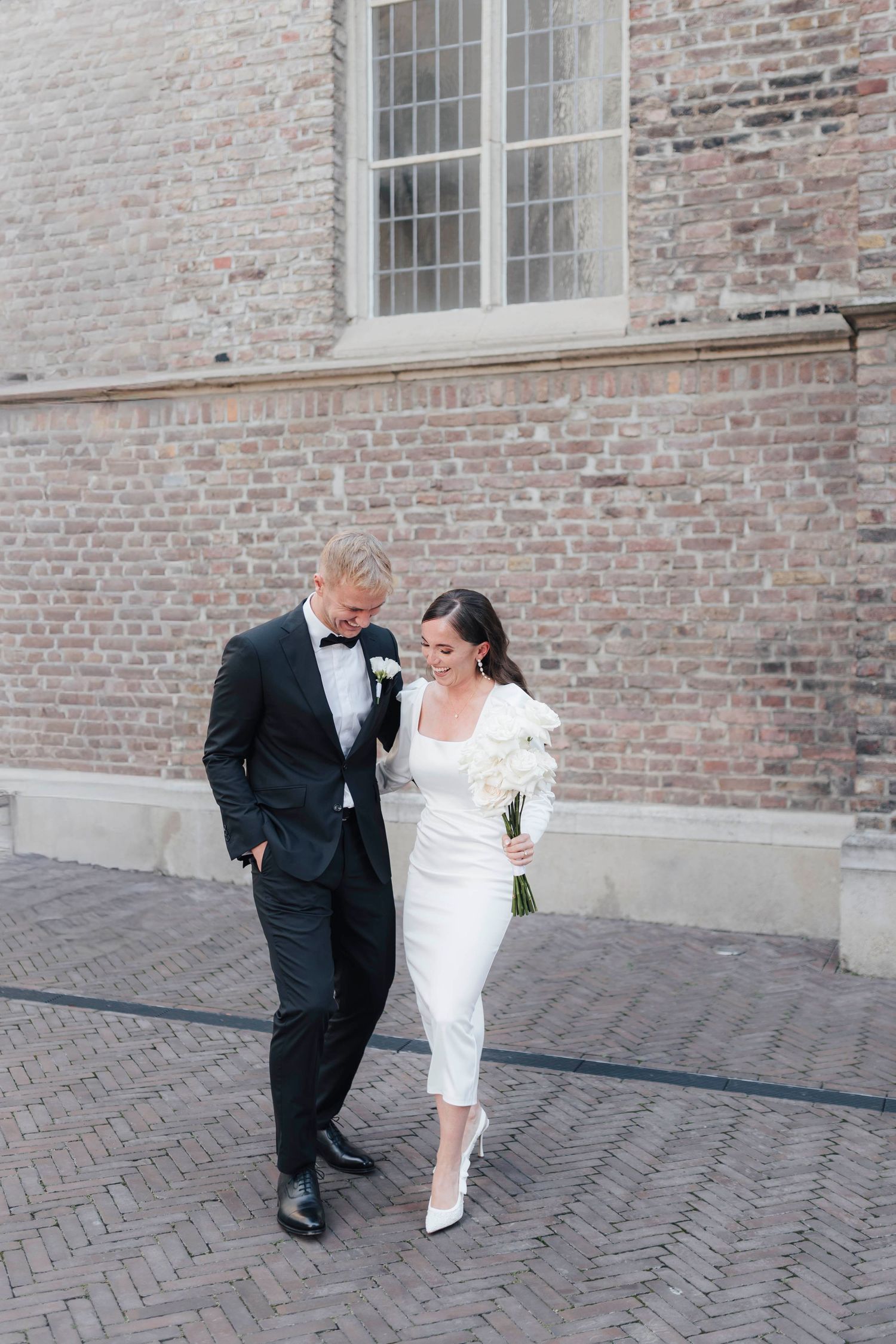 Bride and groom walking together on cobblestone street, she in white midi dress with bouquet, he in black tuxedo.