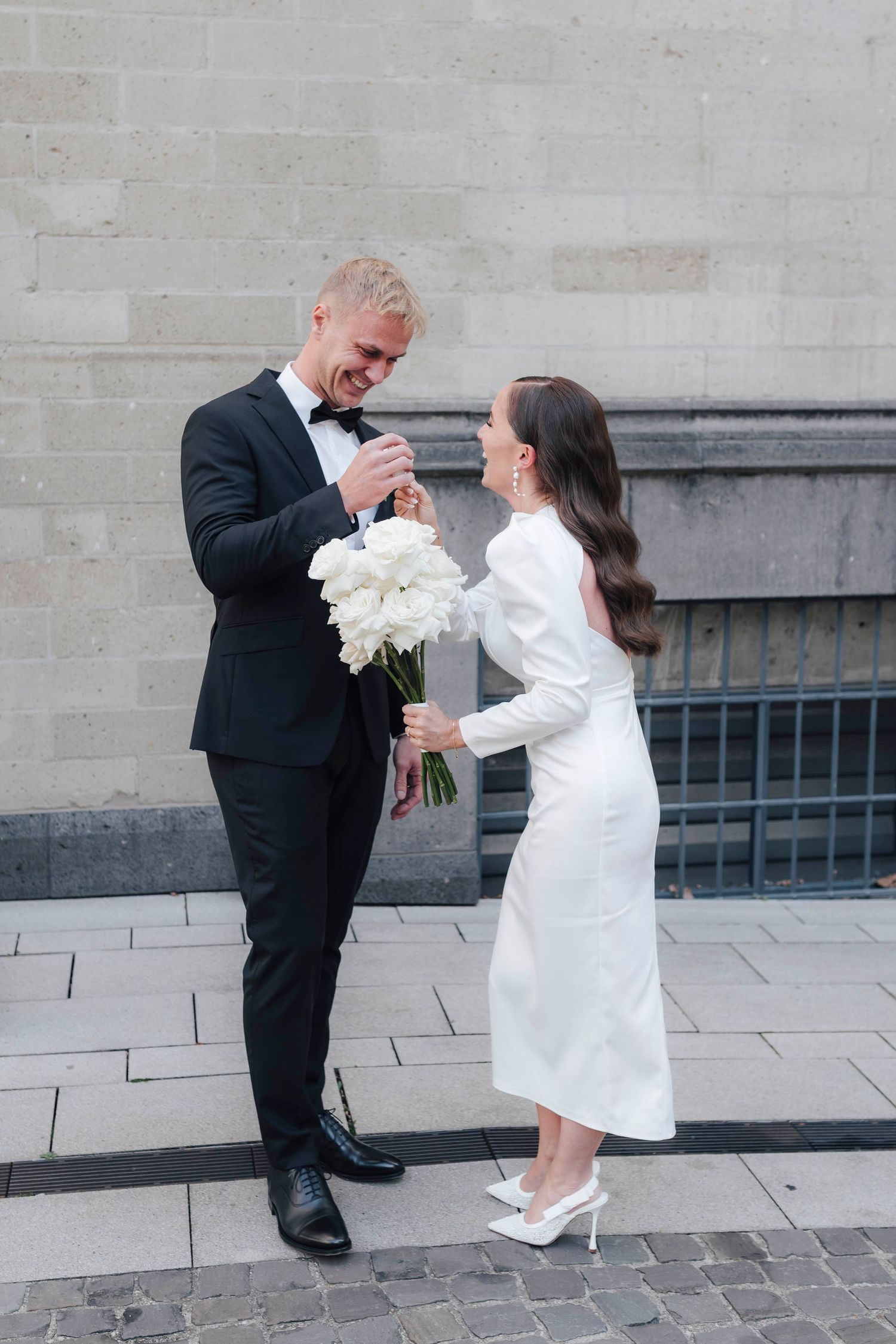 Bride in white midi dress and groom in black tuxedo sharing bouquet of white peonies outdoors.