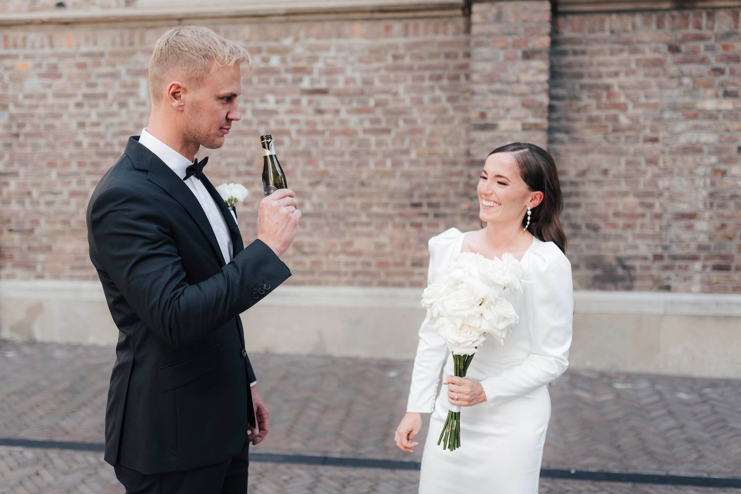 Bride in white gown holding bouquet smiles at groom in black suit opening champagne bottle outdoors.