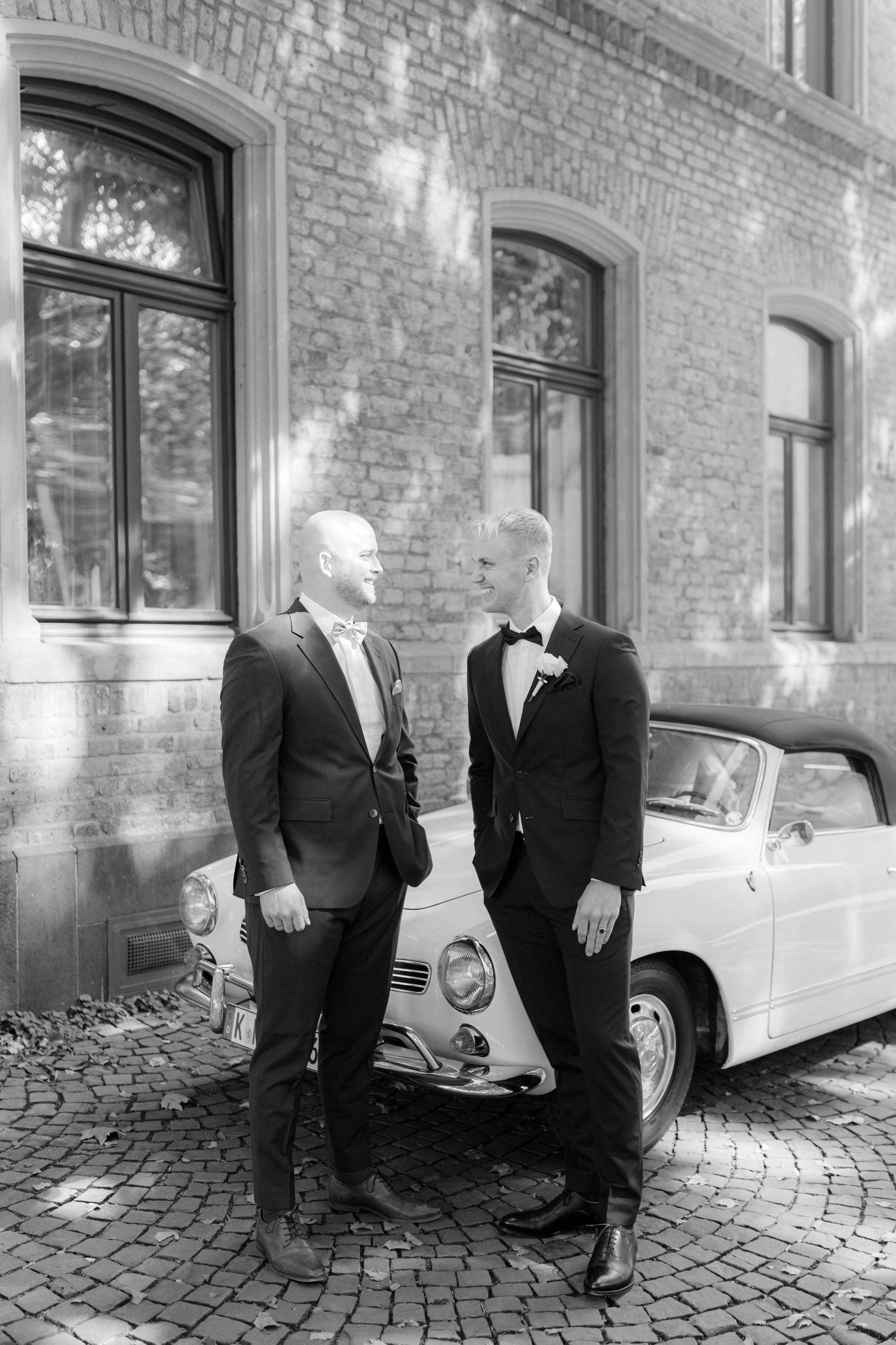 Two grooms in suits stand by a vintage convertible car outside a brick building on their wedding day.