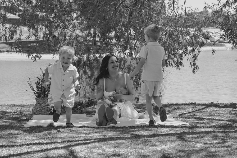 Black and white photo of a mother sitting on a blanket by a lake, smiling with her two young children outdoors.