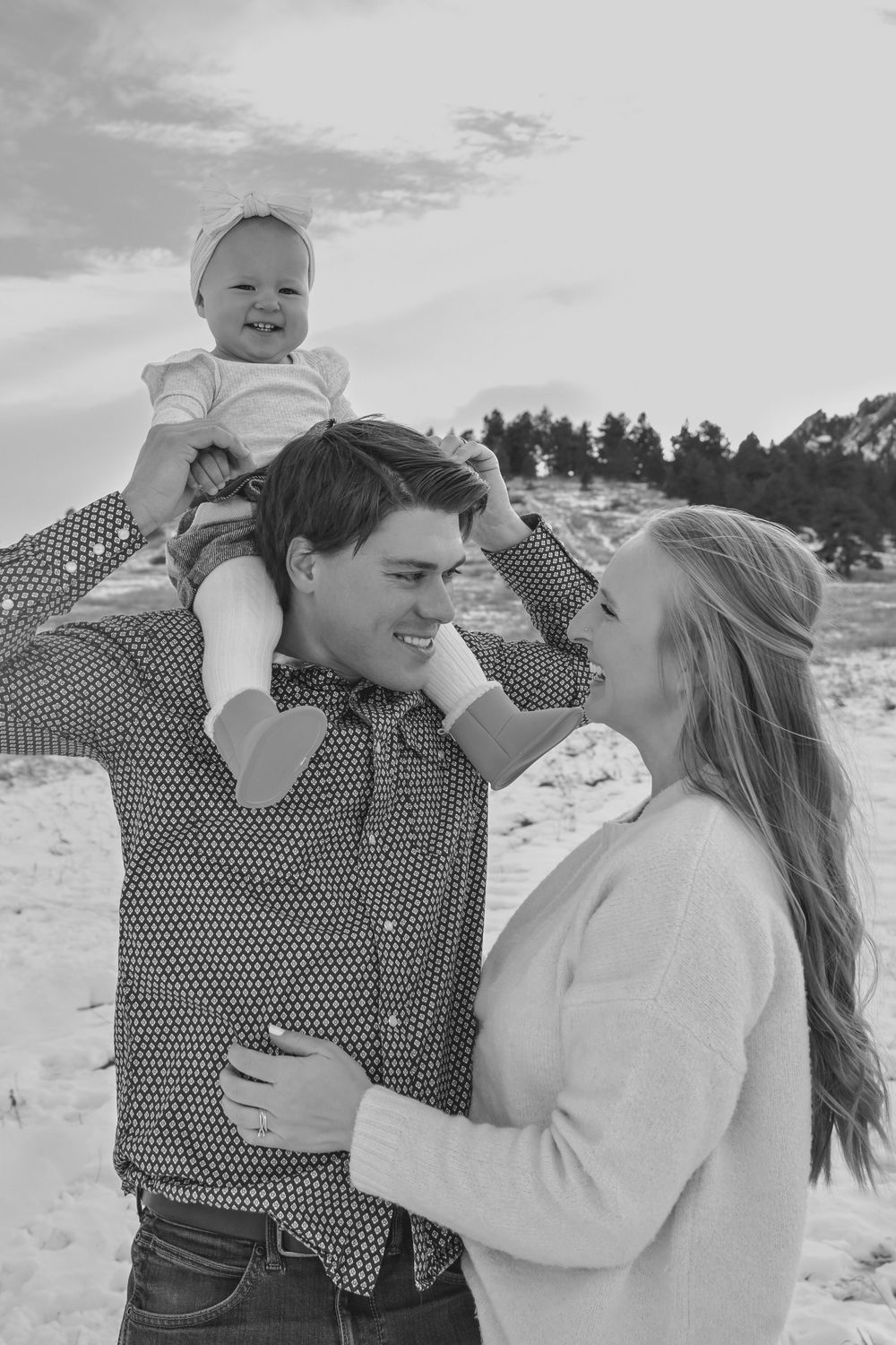 Black and white photo of a happy young family, father holding baby on shoulders while mother smiles lovingly outdoors.