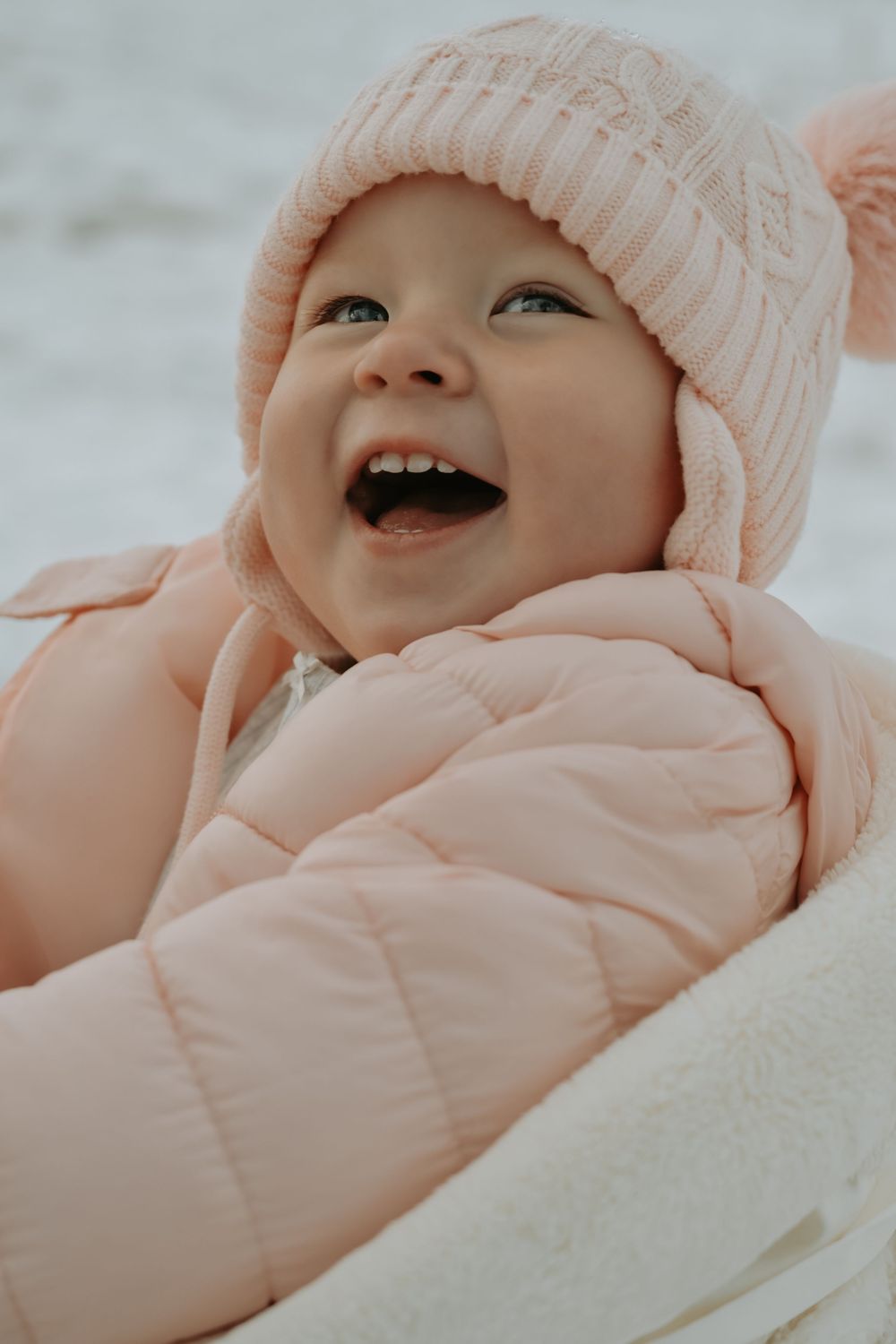 A joyful baby wearing a pink knit hat and puffer jacket laughs outdoors in a snowy winter setting.