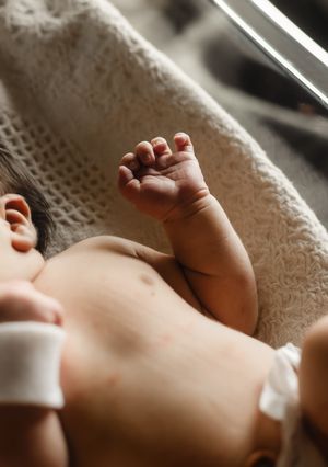 Newborn baby lying on a textured blanket with tiny fist raised, wearing hospital ID bracelet near a window.
