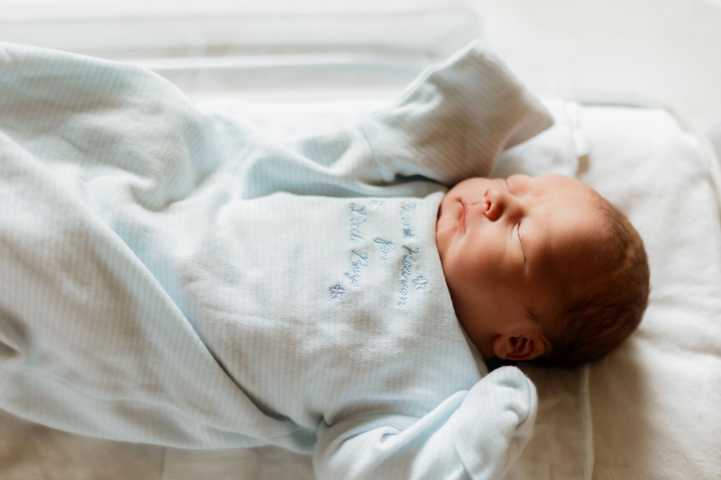 Newborn baby sleeping peacefully wrapped in a soft light blue swaddle blanket in a white bassinet.