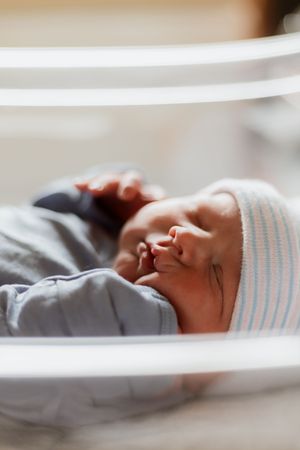 Newborn baby sleeping peacefully in hospital bassinet wearing striped hat and gray outfit.