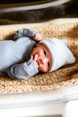 Newborn baby in blue onesie and striped hat lying on beige textured blanket in hospital bassinet.