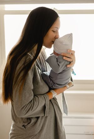 A new mother in a gray robe tenderly kisses her swaddled newborn baby wearing a white knit hat near a bright window.