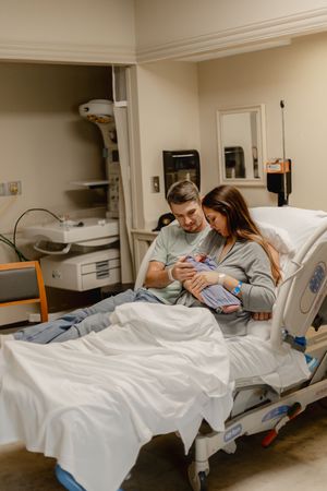 New parents in hospital room holding their newborn baby together on a medical bed after delivery.
