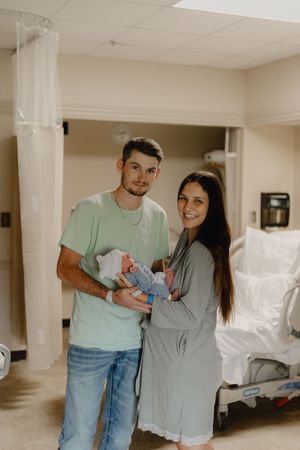 New parents smiling in hospital room, father holding newborn baby, mother in gray robe beside him.
