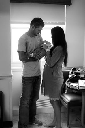 Black and white photo of new parents holding their newborn baby in a hospital room near a window.