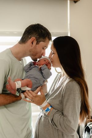 New parents sharing a tender kiss over their newborn baby in a hospital room, both wearing patient wristbands.