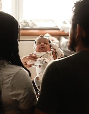 Parents gazing lovingly at their sleeping newborn baby dressed in a white onesie in a warmly lit room.