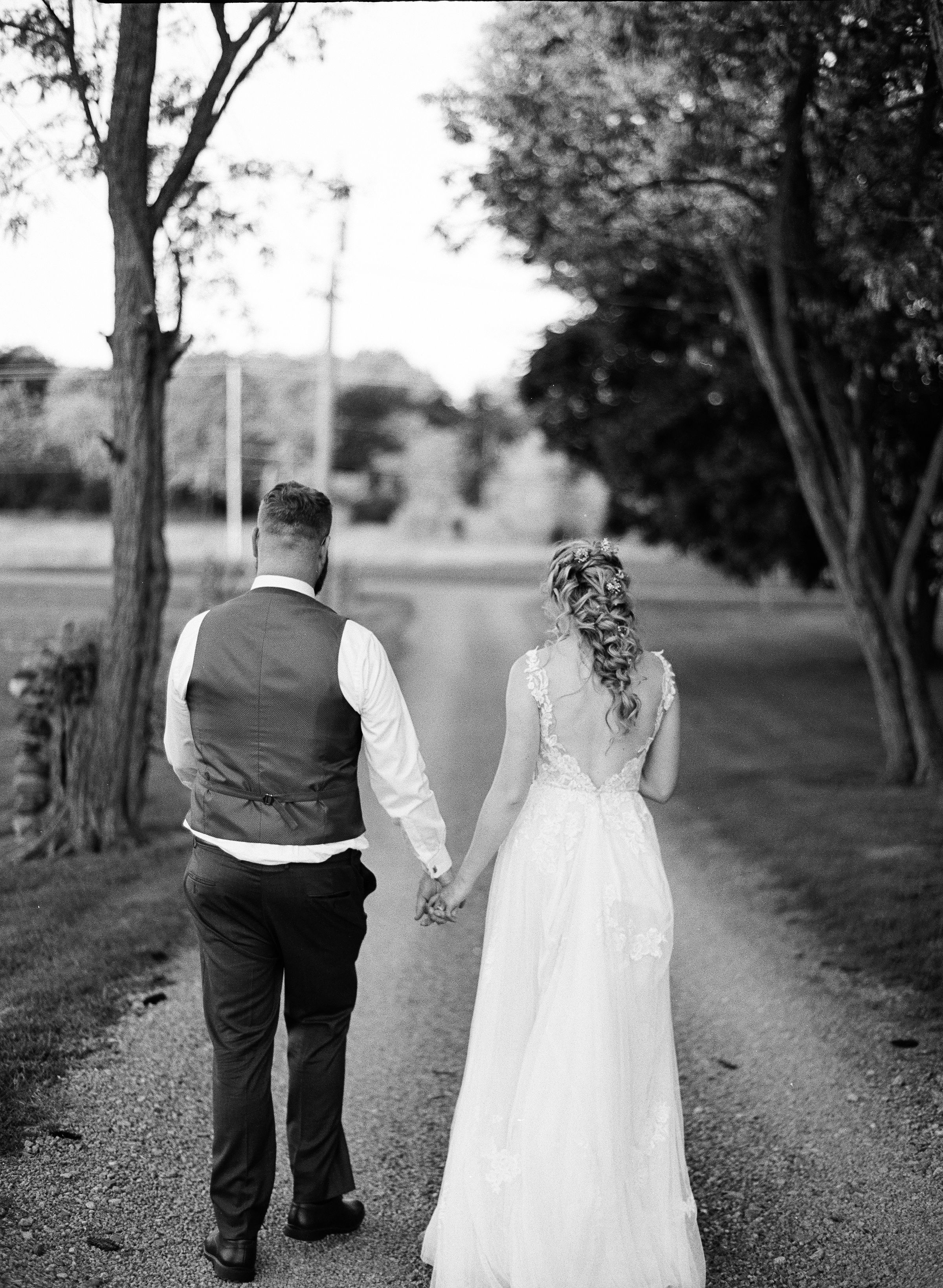 Black and white photo of a bride and groom walking hand in hand down a tree-lined path on their wedding day.