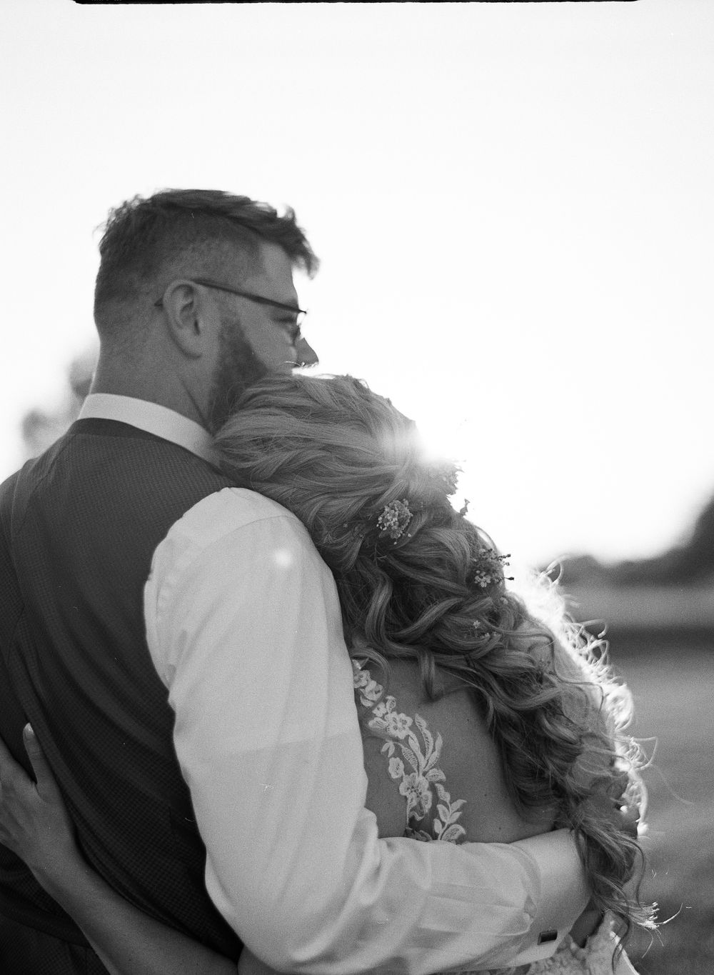 Black and white photo of a couple embracing at their wedding, backlit by golden sunlight in an outdoor setting.
