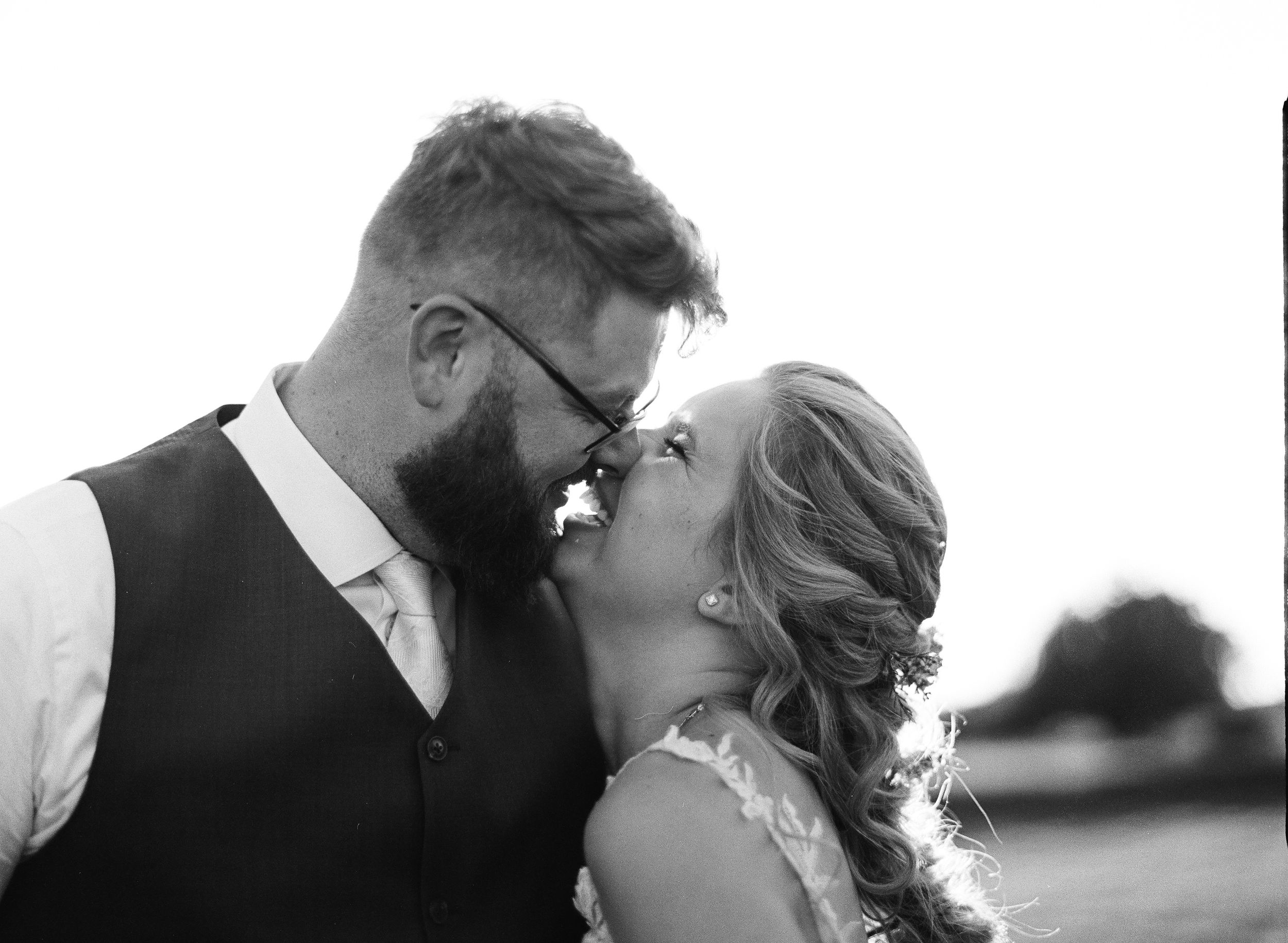 Black and white wedding photo of a bearded groom and smiling bride sharing a romantic kiss outdoors.