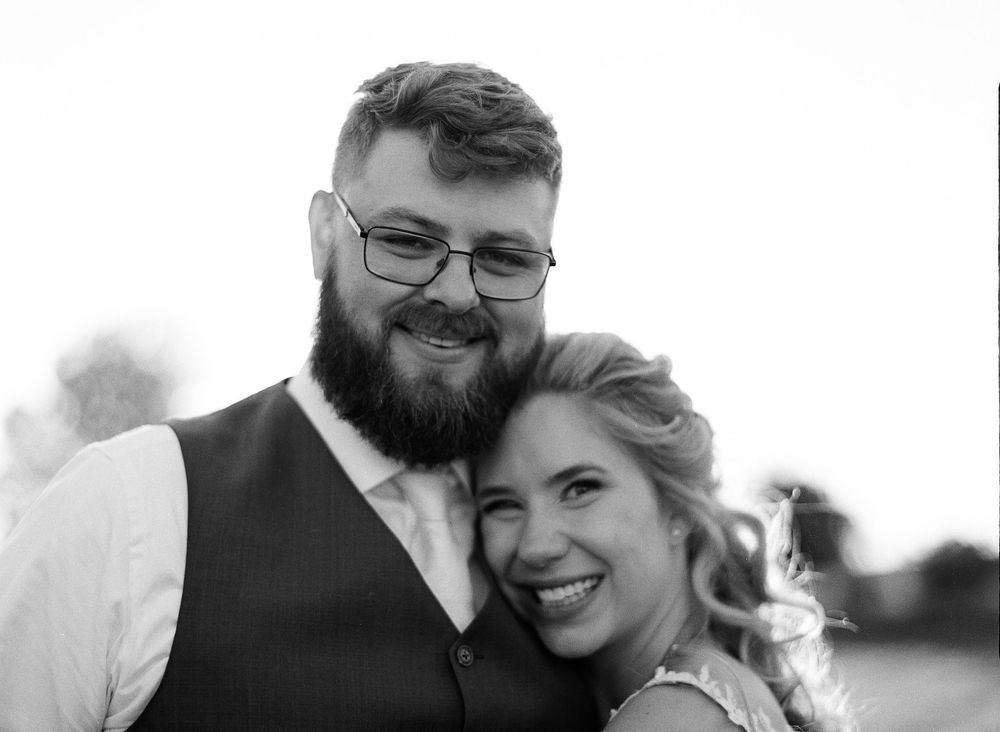 Black and white wedding photo of smiling couple, bearded groom in vest and bride with updo hairstyle embracing outdoors.