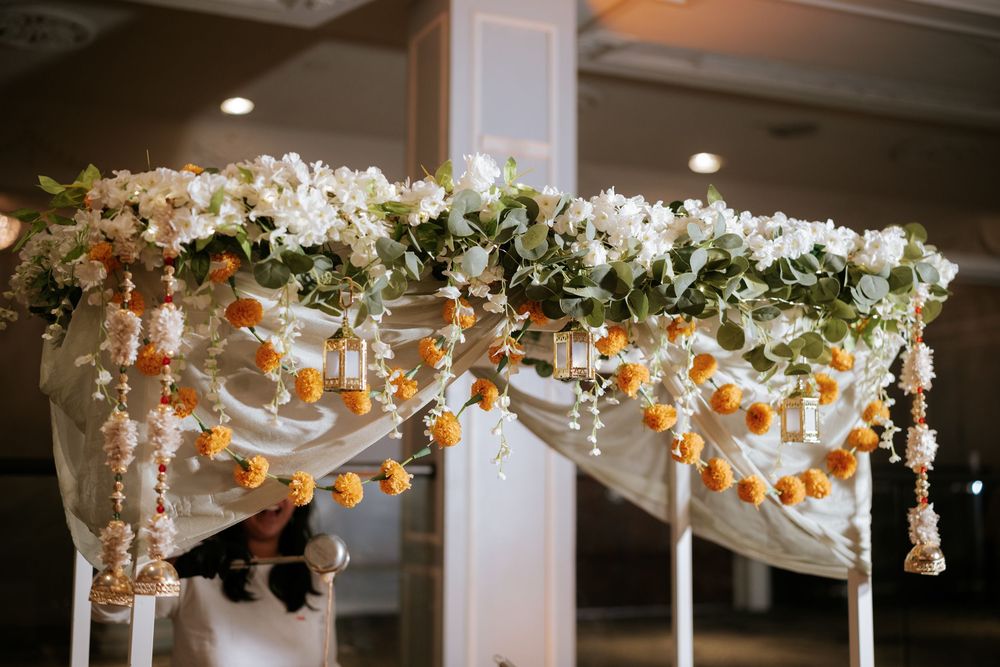 Elegant wedding mandap decorated with white orchids, green garlands, and hanging orange marigold strings in a banquet hall.