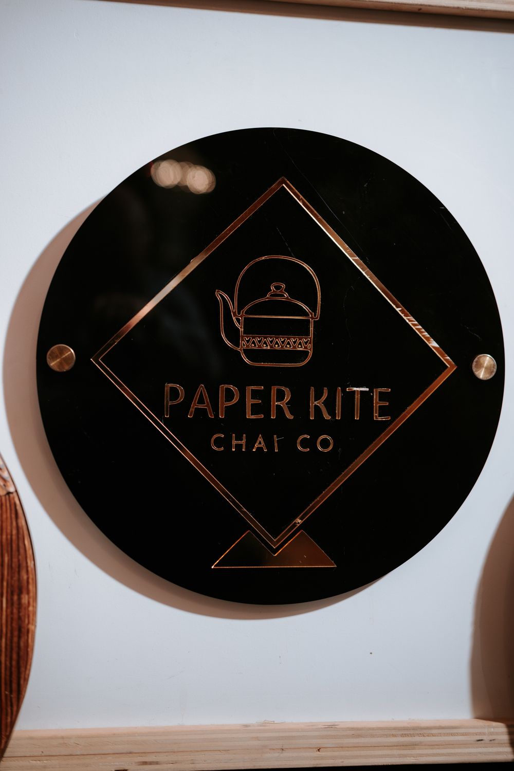 Black circular sign with rose gold logo and text for Paper Kite Chai Co featuring a teapot illustration.