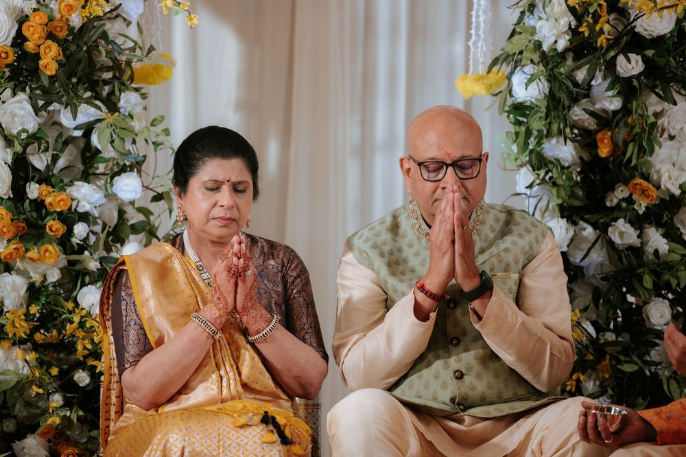 Elderly Indian couple in traditional attire performing Namaste prayer gesture at a floral decorated ceremony.