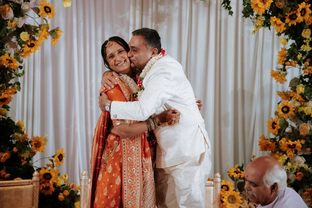 Couple embracing at Indian wedding ceremony, bride in orange lehenga, groom in white sherwani, sunflower floral backdrop.