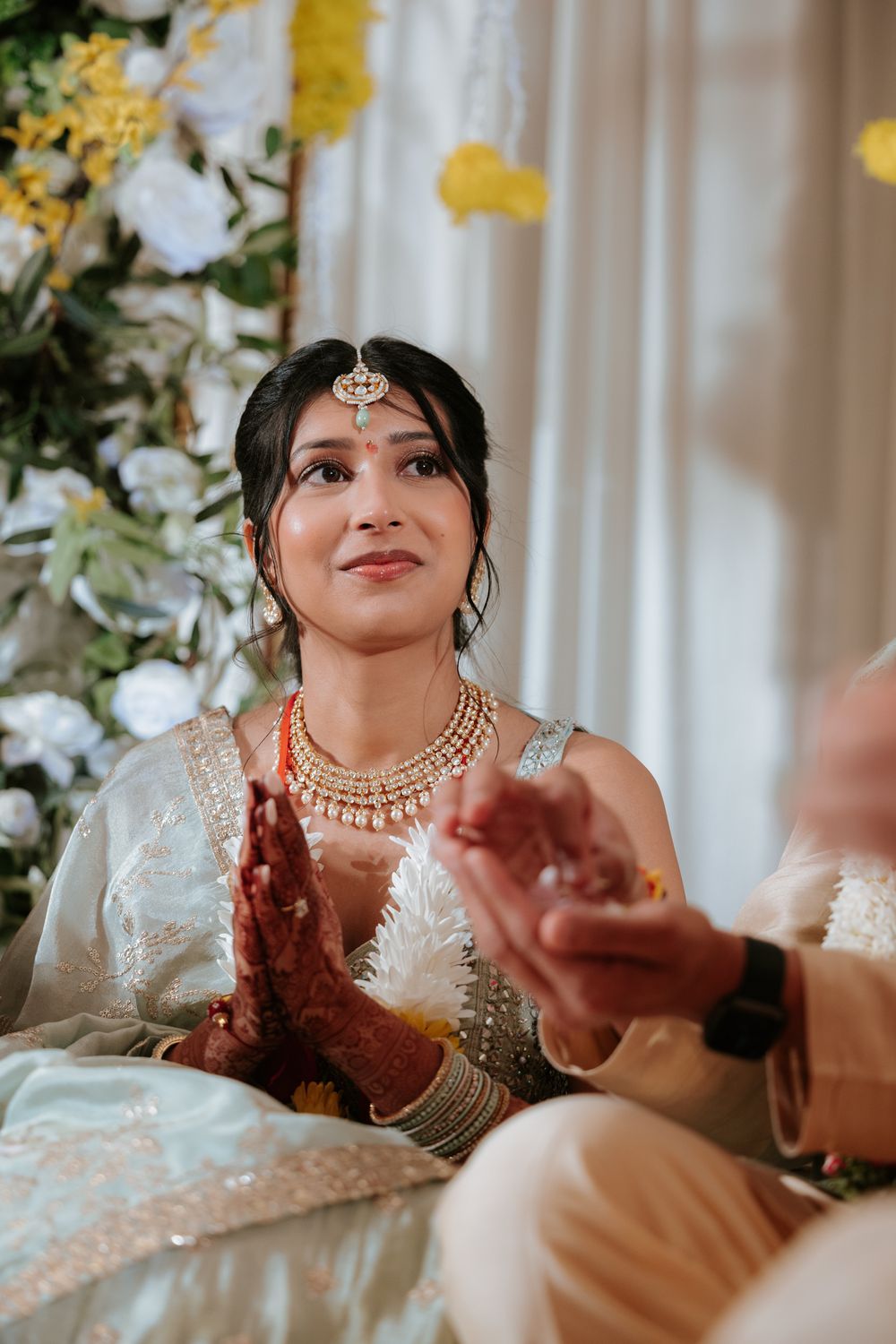 Indian bride in white lehenga with gold jewelry and mehndi hands joined in prayer during wedding ceremony.