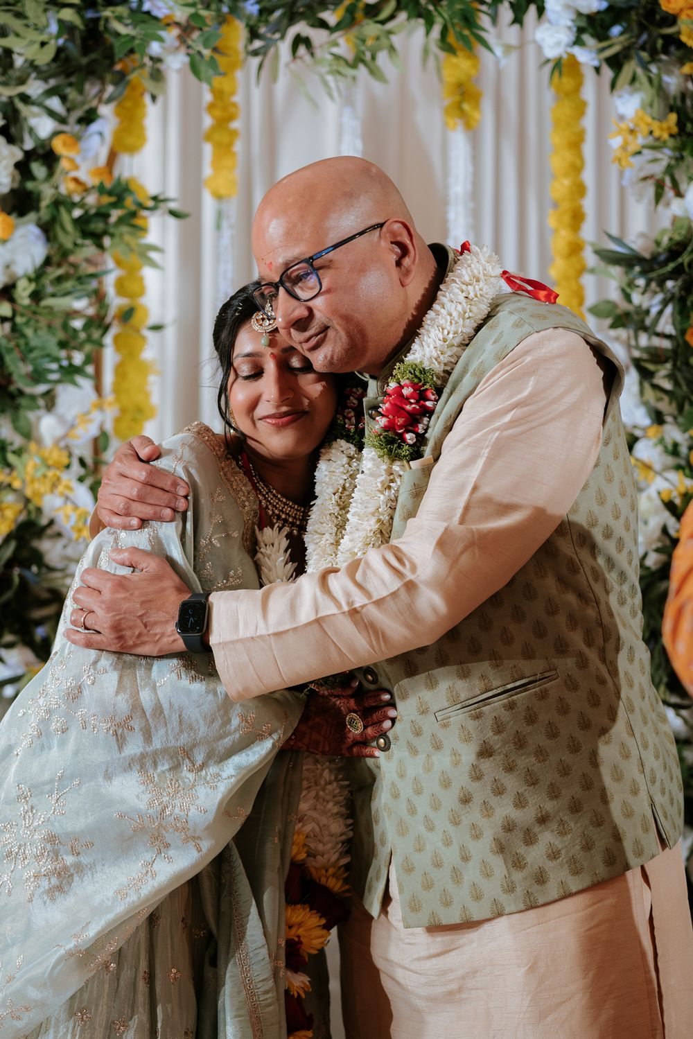 Emotional embrace between bride and groom in traditional Indian wedding attire with floral garlands and yellow marigold backdrop.