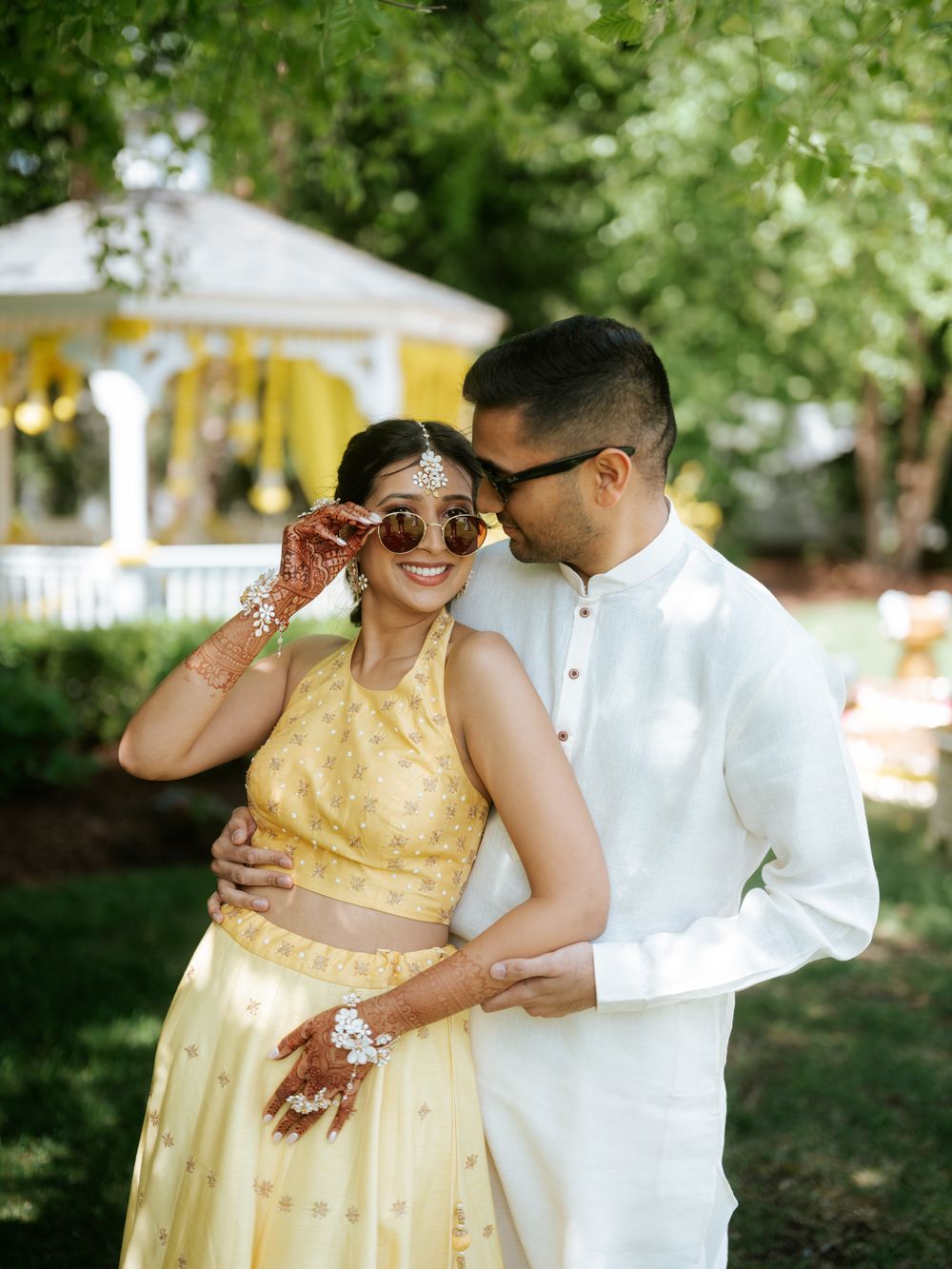 Indian couple in yellow lehenga and white kurta sharing a romantic moment in a garden with gazebo.