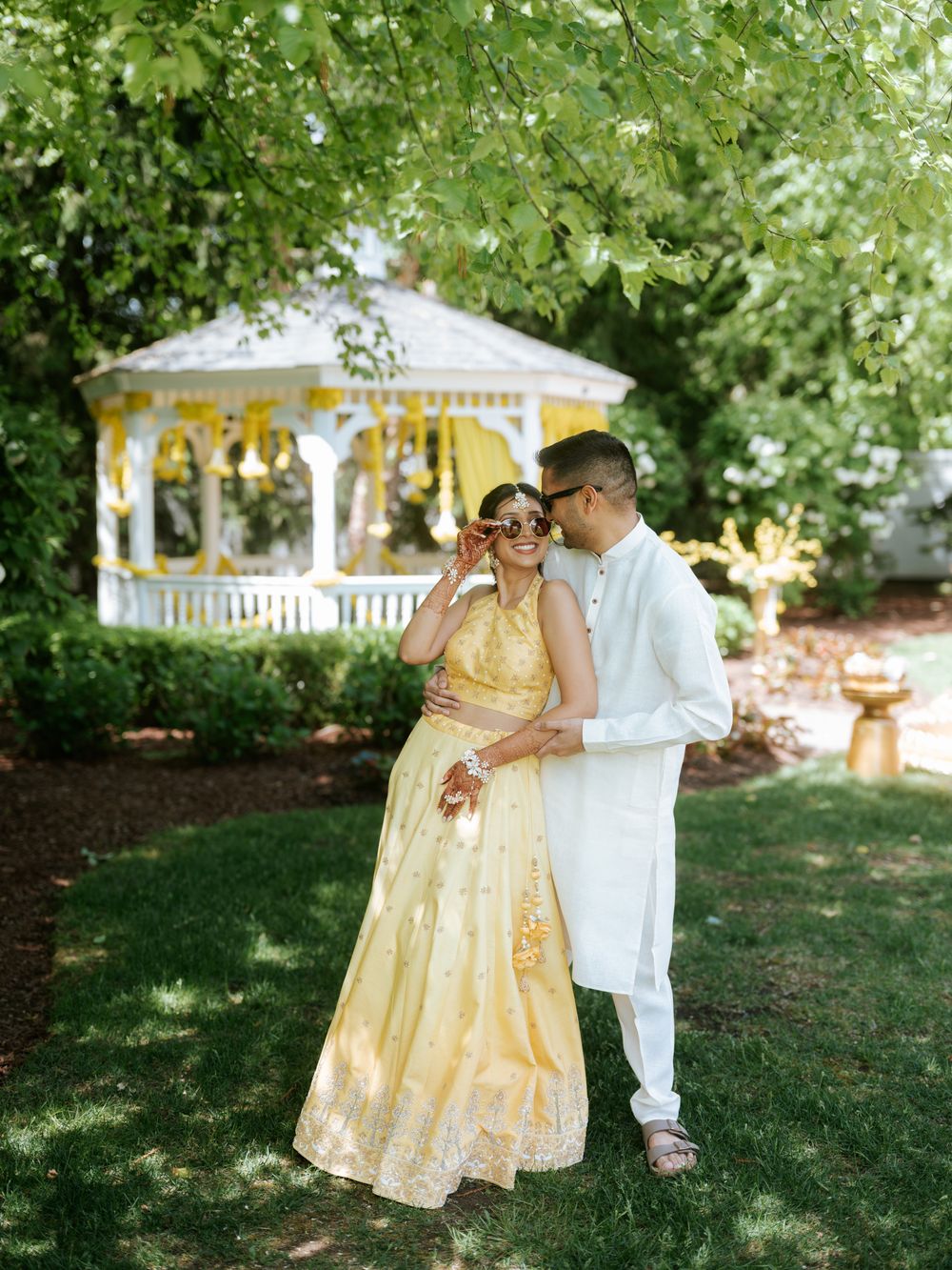 Couple in Indian wedding attire laughing together in lush garden with white gazebo decorated with yellow flowers.