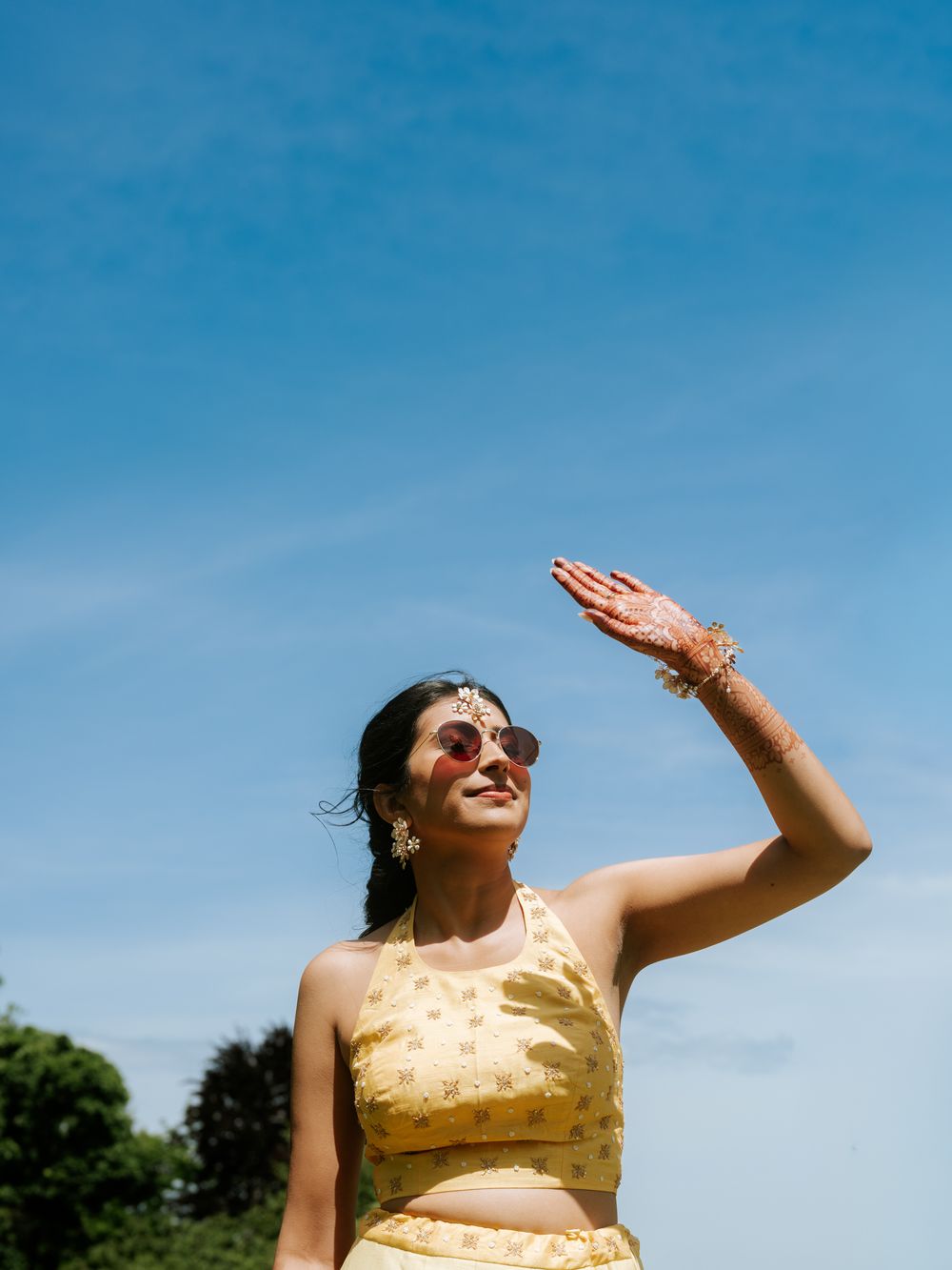 Bride in yellow lehenga with henna hands shielding eyes under bright blue sky during outdoor ceremony.