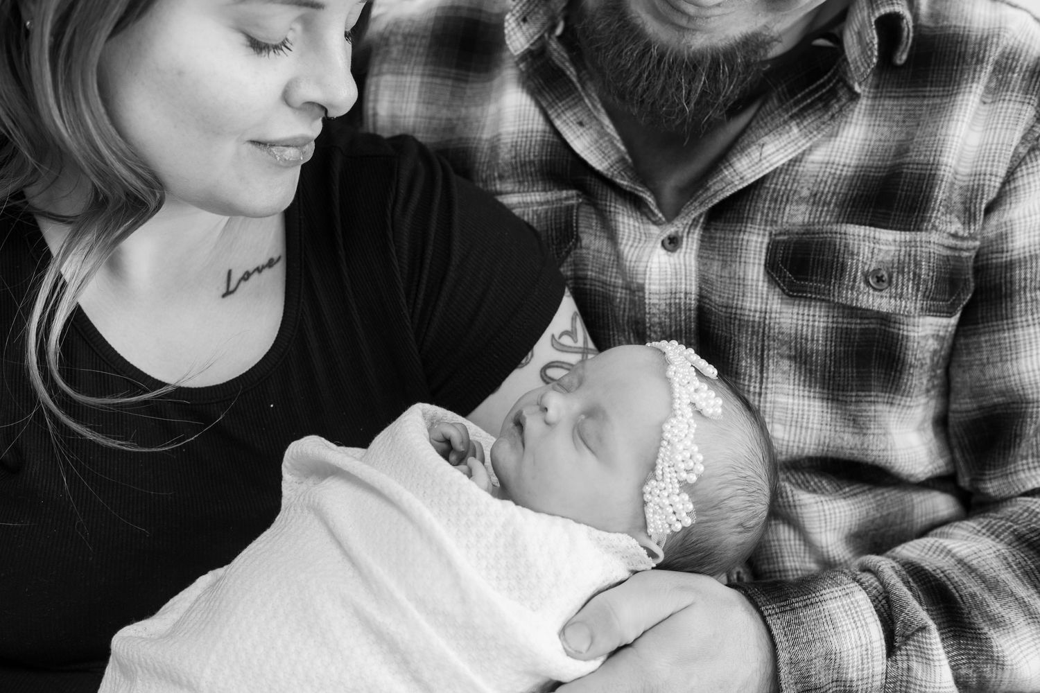Black and white photo of new parents lovingly gazing at their swaddled newborn baby girl wearing a floral headband.