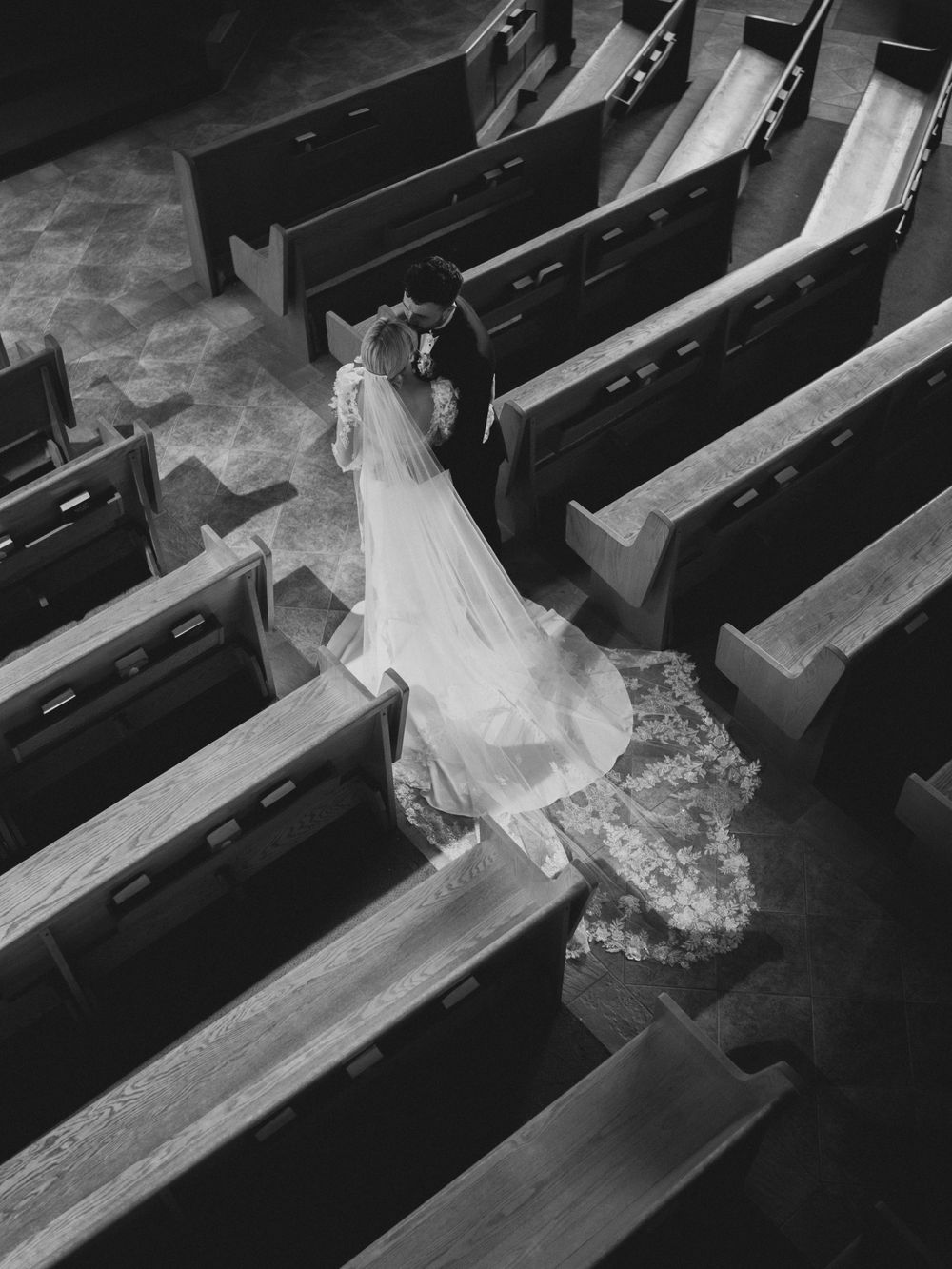 Black and white aerial view of bride and groom embracing in church aisle surrounded by wooden pews.