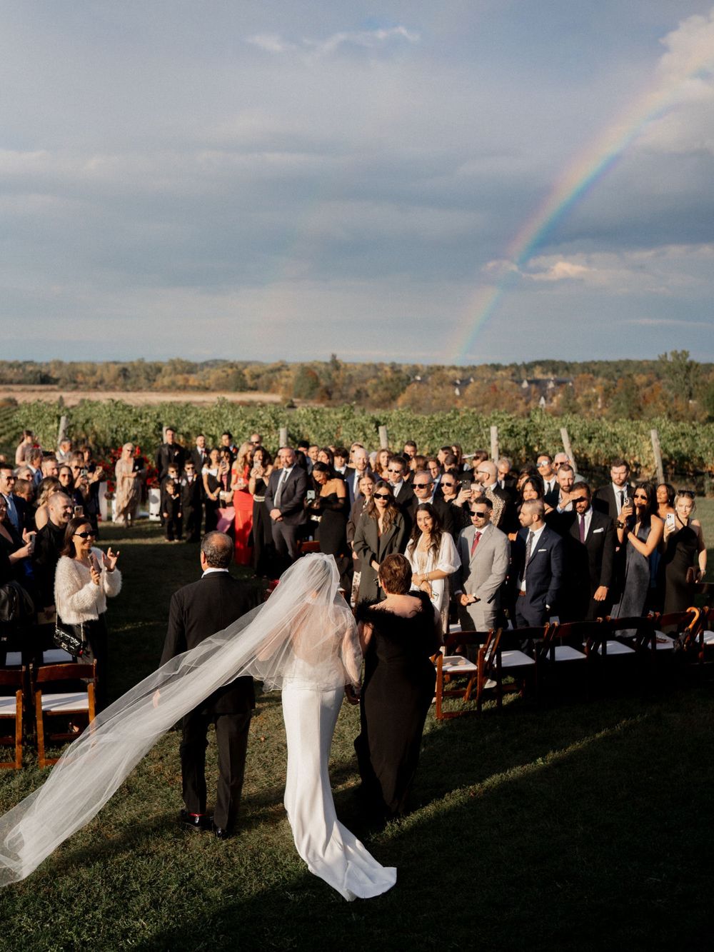 Bride and groom walk down outdoor aisle at vineyard wedding ceremony under a stunning rainbow.