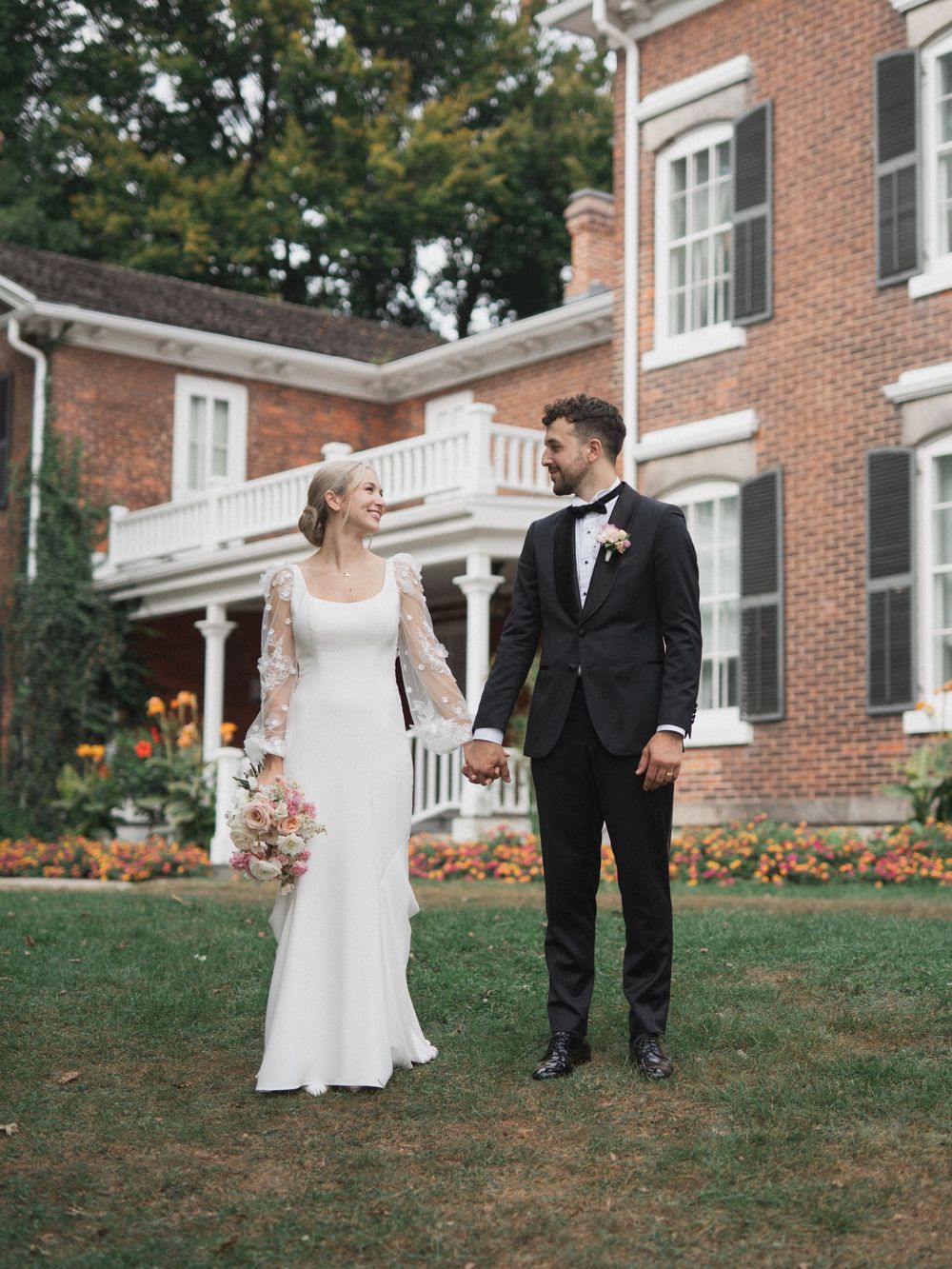 A bride and groom hold hands on a lawn in front of a brick manor, she in lace sleeves, he in a black tuxedo.