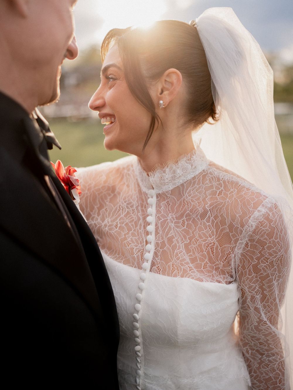 Smiling bride in lace high-neck wedding dress and veil stands with groom in black suit outdoors in golden sunlight.