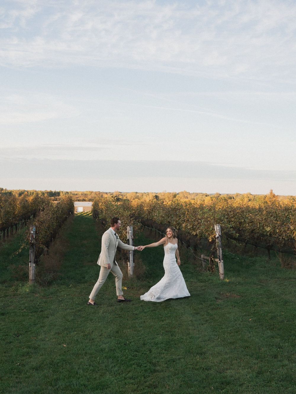 Bride and groom holding hands dancing in a scenic vineyard at golden hour.