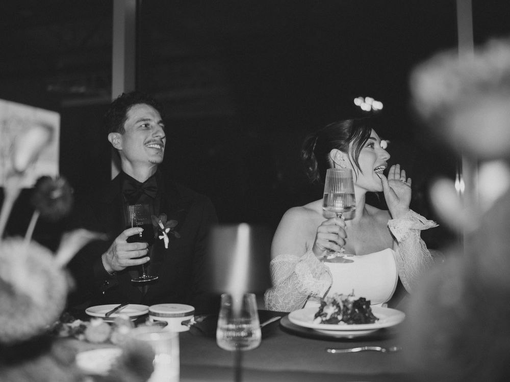 Black and white photo of bride and groom laughing at wedding reception dinner table with champagne glasses.