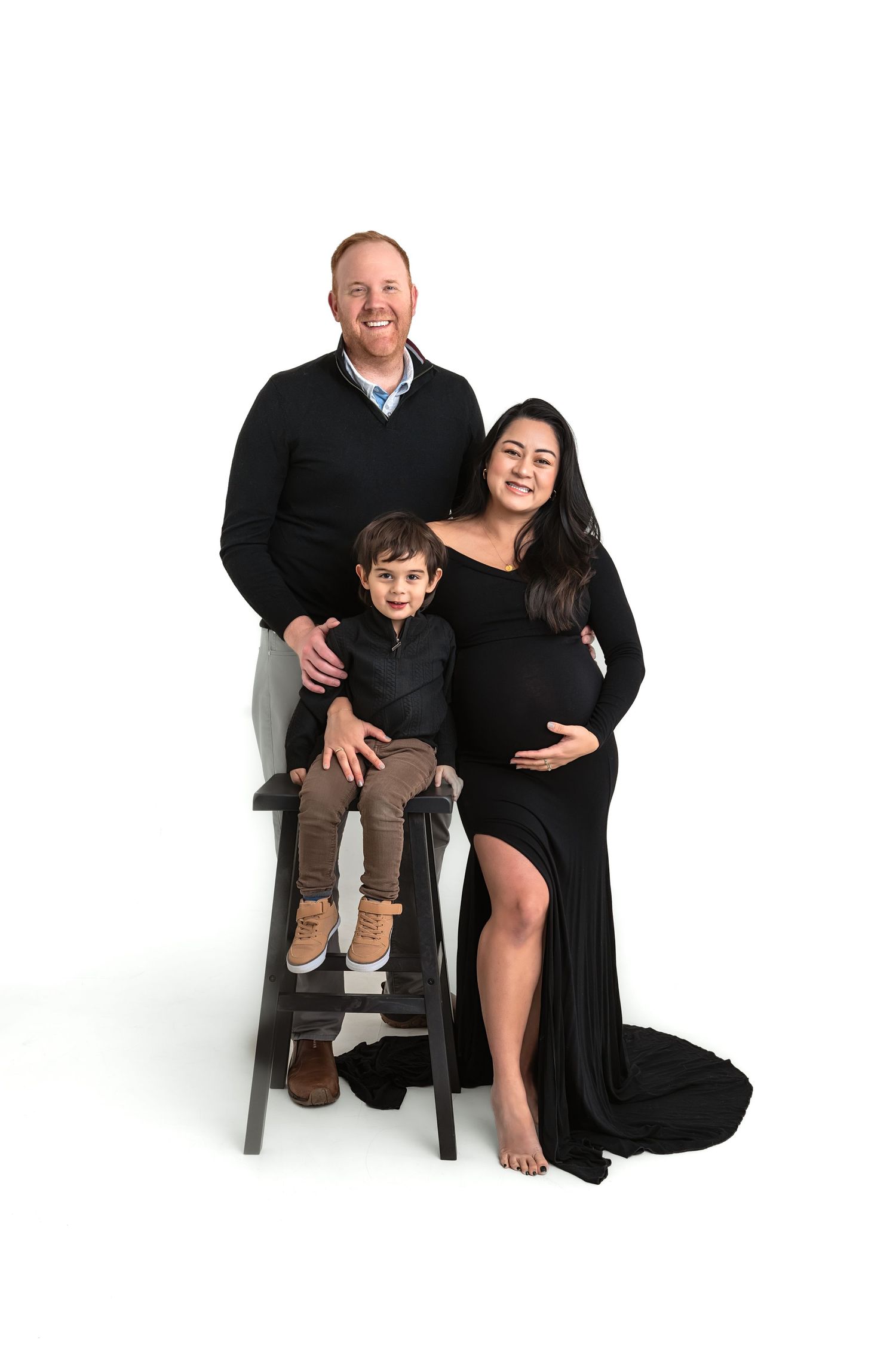 Maternity photo of pregnant woman in black gown with husband and young son on white studio background.