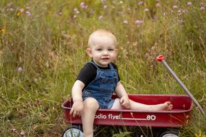 Smiling baby in denim overalls sitting in a red Radio Flyer wagon in a wildflower meadow.