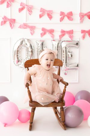 Baby girl in pink dress sits in wooden rocking chair at first birthday party with balloons and ONE balloon letters.