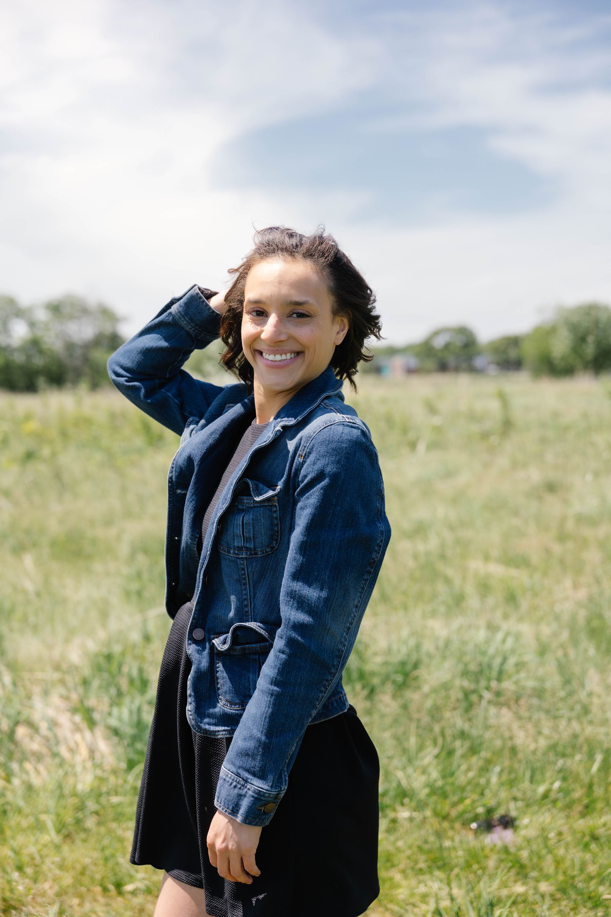 Smiling young woman in denim jacket and black skirt standing in green field on sunny day.