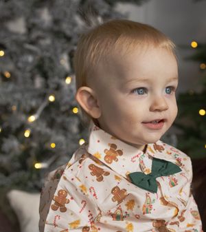 Smiling baby in gingerbread print outfit with green bow tie in front of Christmas tree with warm lights.