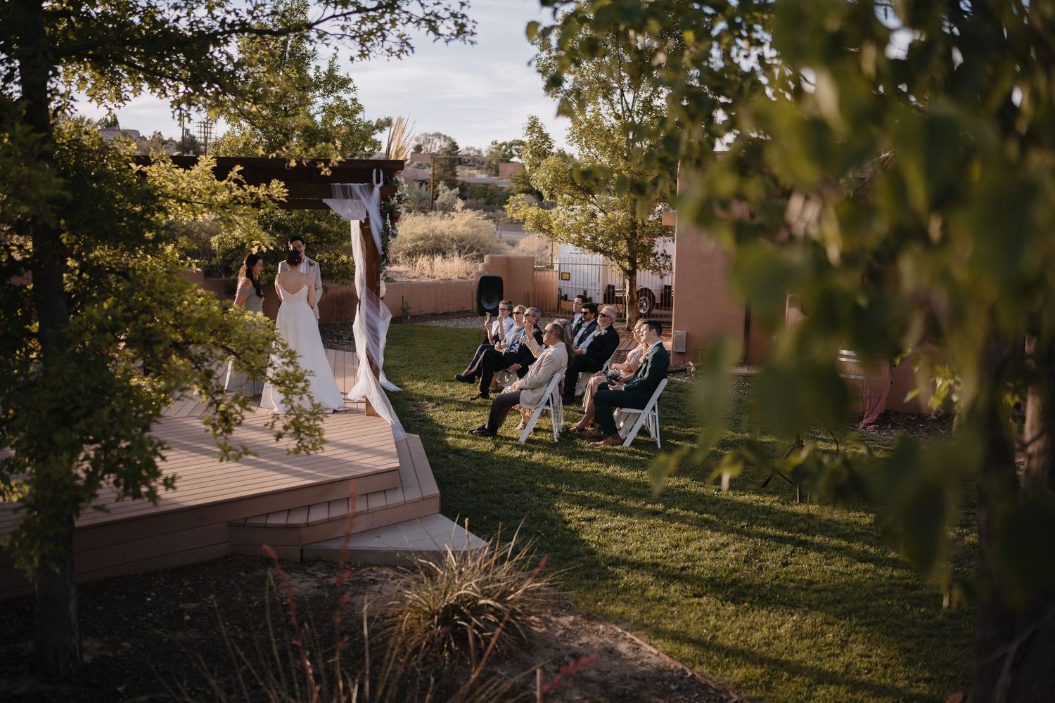 Intimate outdoor wedding ceremony with guests seated on white chairs surrounded by lush green trees.