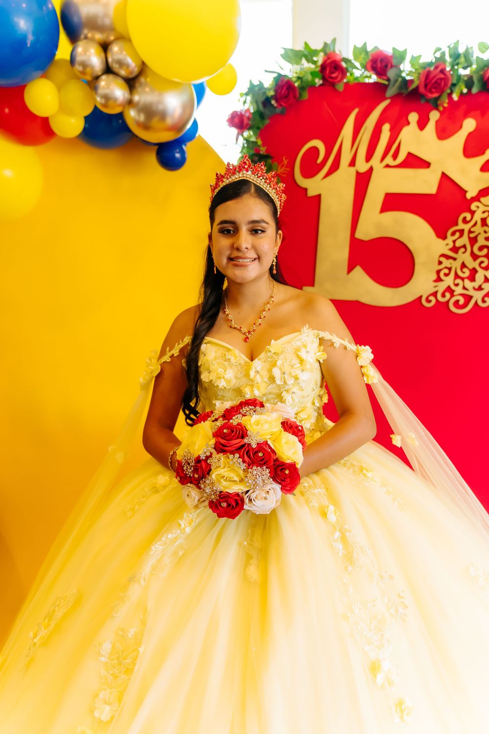 Young Latina girl in yellow quinceañera ball gown with red roses bouquet and tiara poses at her 15th birthday celebration.