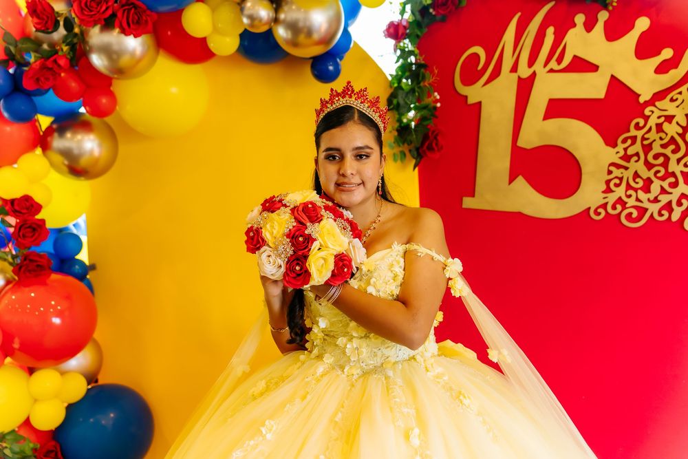 A young girl in a yellow quinceañera gown holds a red and white bouquet at her Mis 15 celebration with colorful balloons.