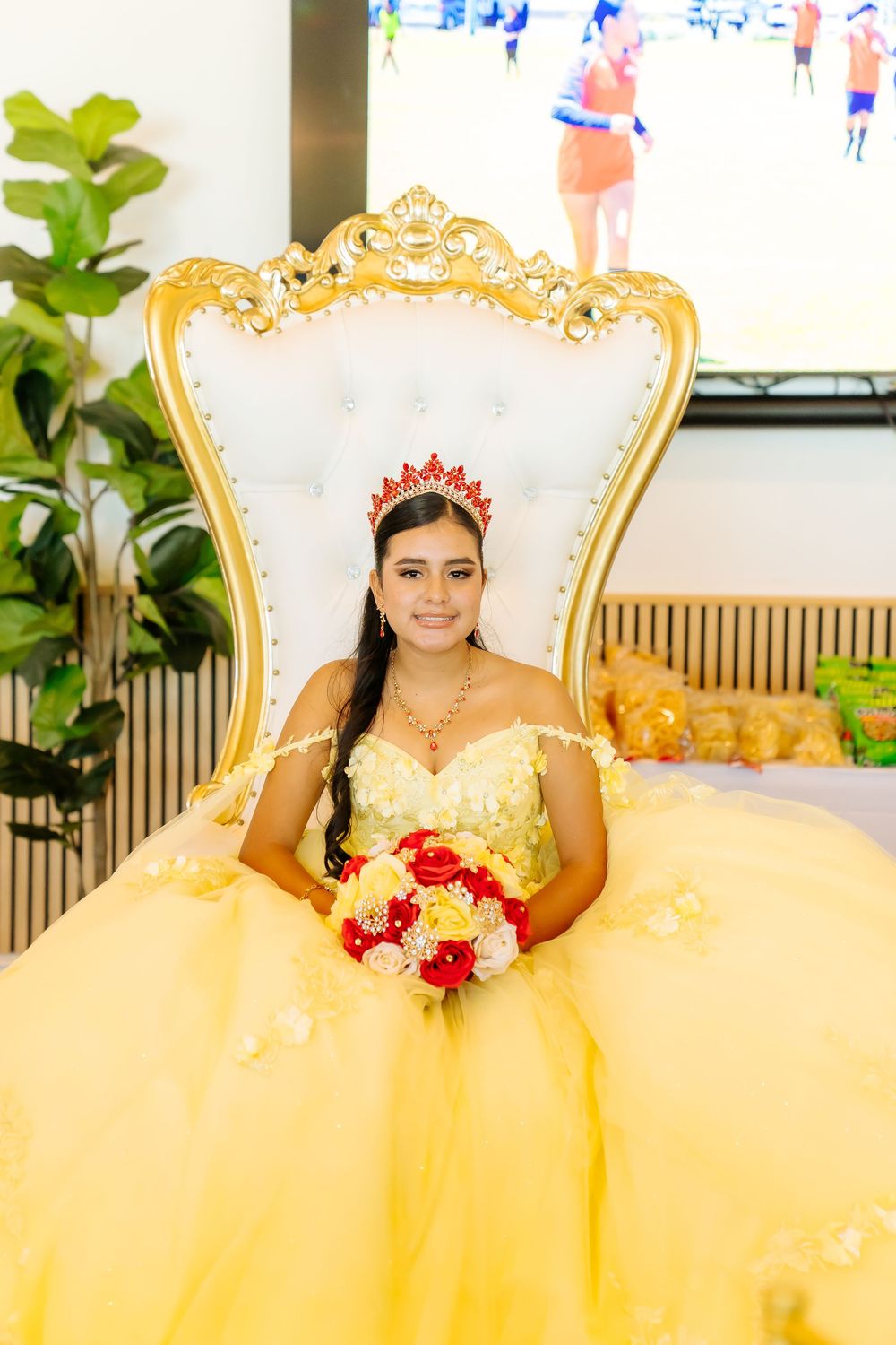Young Latina girl in yellow quinceañera gown with red crown holds bouquet, seated on ornate white and gold throne chair.