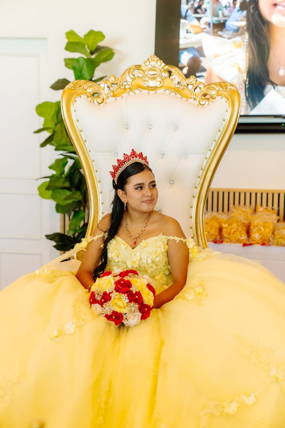 Young woman in yellow quinceañera gown with red crown holds floral bouquet seated on ornate gold throne chair.