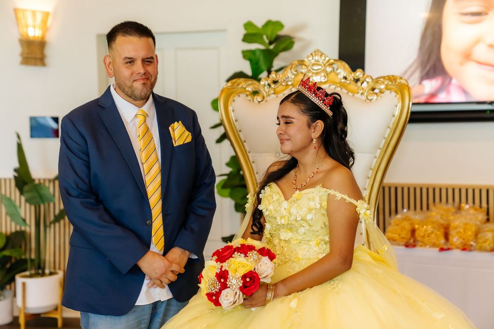 A girl in a yellow quinceañera gown holds a bouquet beside a man in a blue suit at a elegant celebration.