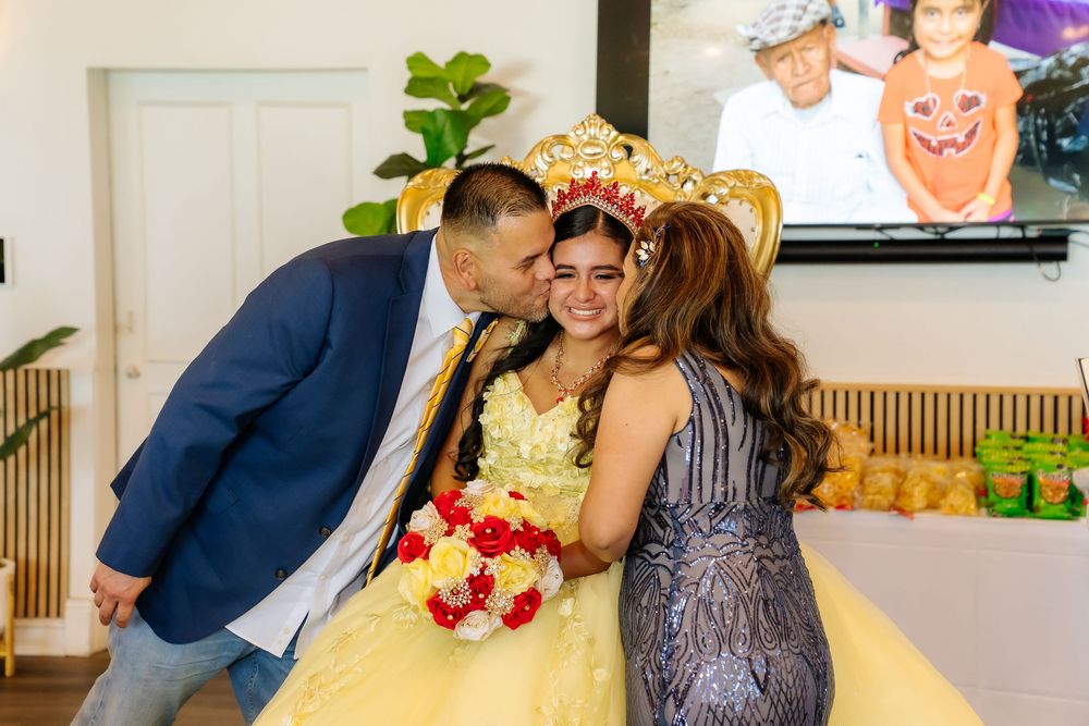 A young girl in a yellow quinceañera gown and crown receives kisses from her parents at her celebration party.