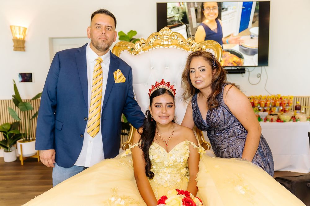 A young girl in a golden quinceañera gown and tiara poses with a man in a blue suit and a woman in a gray dress.